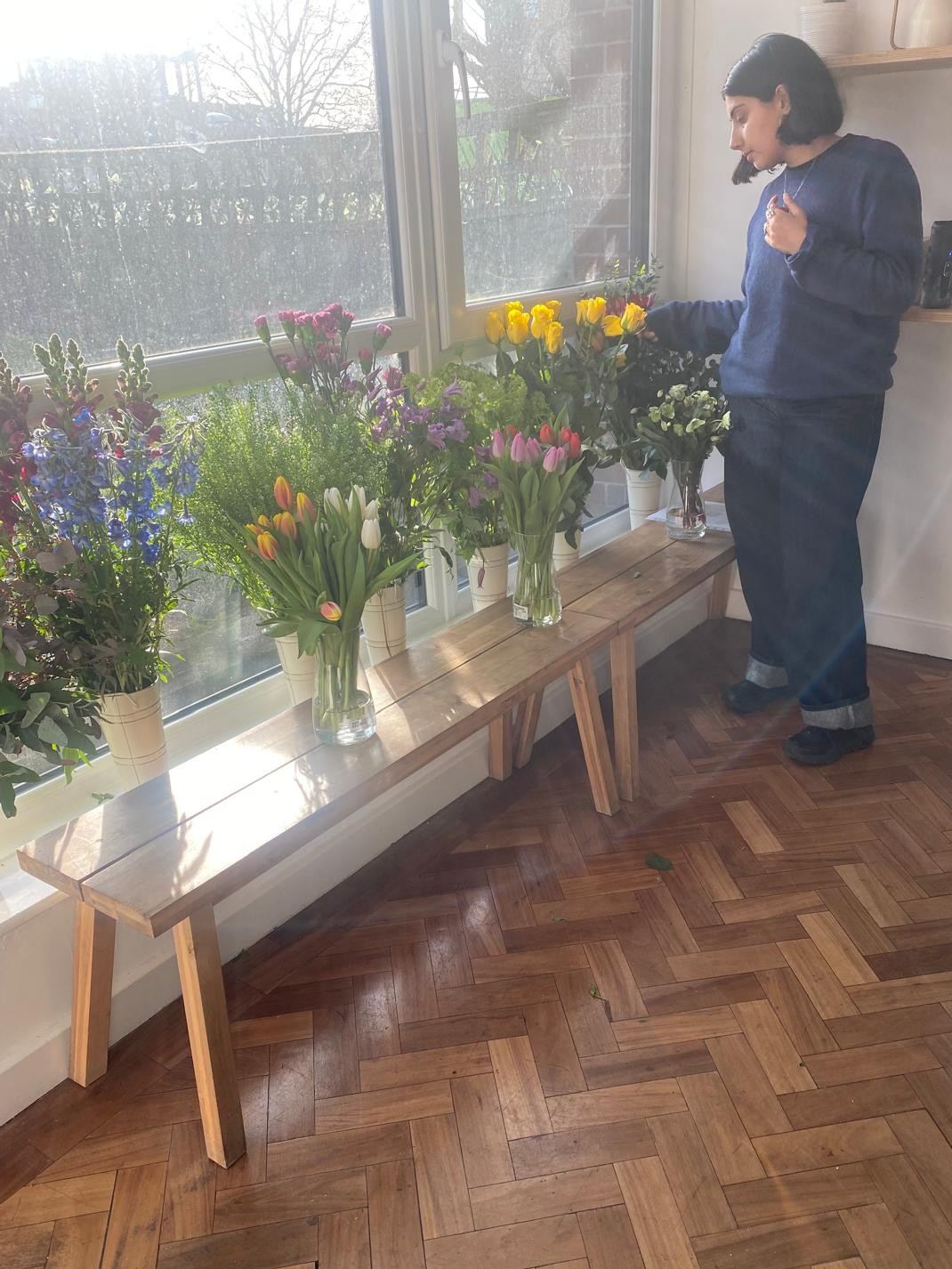 Choosing stems from vases of tulips, delphiniums and stocks along a sunny window bench